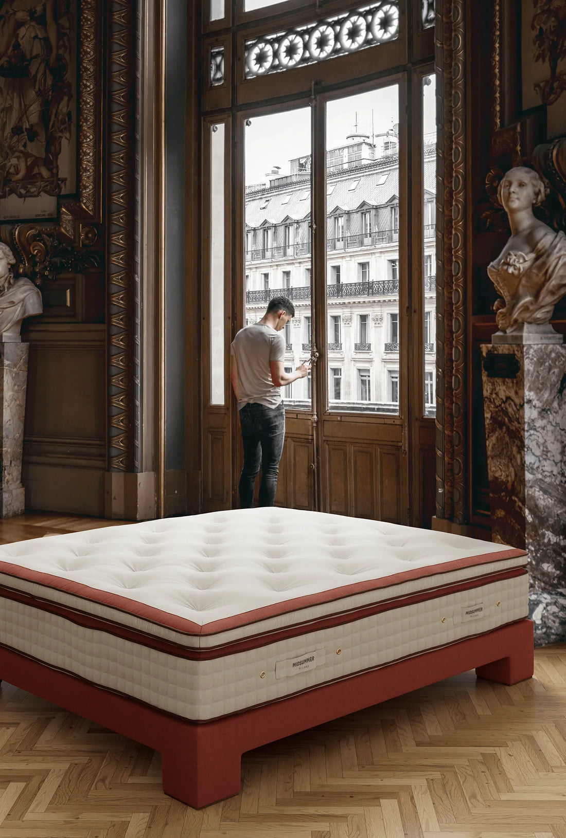 Luxury Midsummer-Milano mattress in an elegant, historic European room with parquet floors and classical sculptures, with a man standing by a large ornate window overlooking a Parisian building.