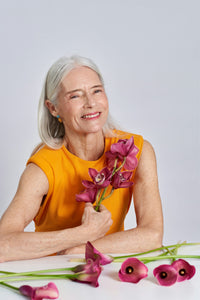 Smiling older woman in orange dress holding purple flowers against a light background.