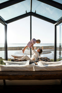 A mother lifts her baby in the air while relaxing on a luxury bed in a glass-walled room.