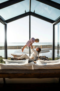 Mother and child playing on a bed in a glass room with a scenic coastal view