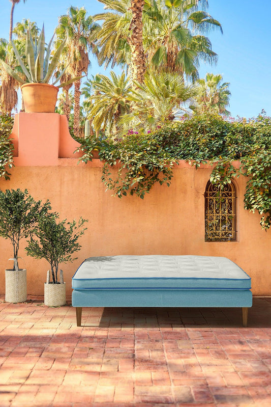 Luxury upholstered bed with white mattress outdoors by terracotta wall, palm trees in background