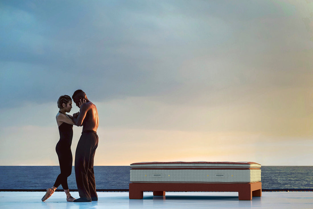 A couple dancing beside a luxury mattress on a terrace overlooking the ocean at sunset.
