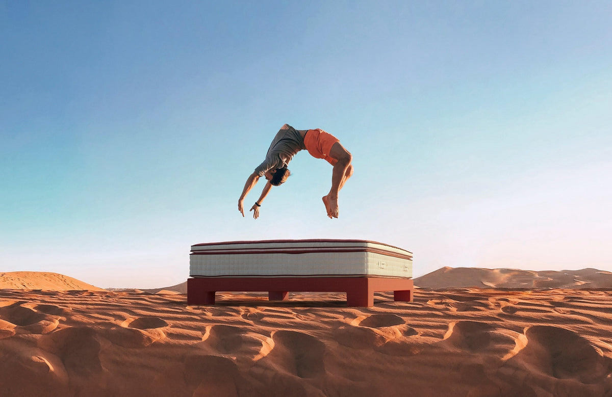 Man doing backflip onto luxury mattress in desert landscape under clear sky
