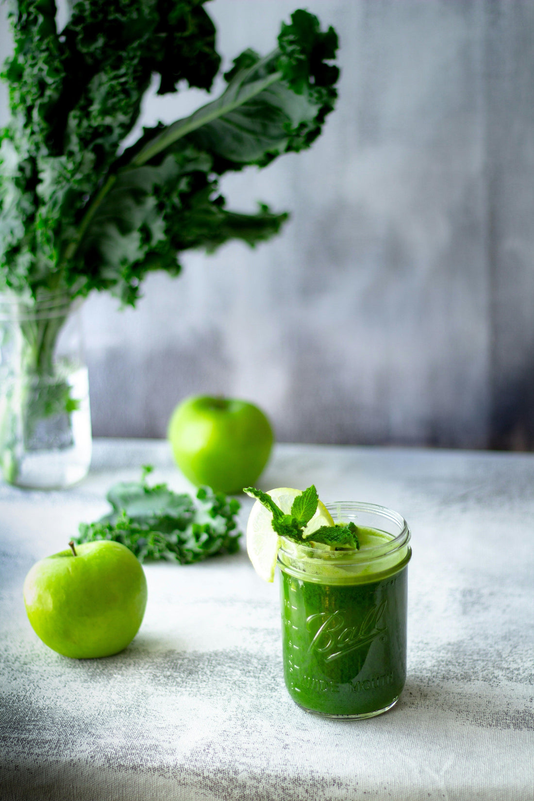 Green smoothie in glass jar with lime and mint, surrounded by kale and green apples on table