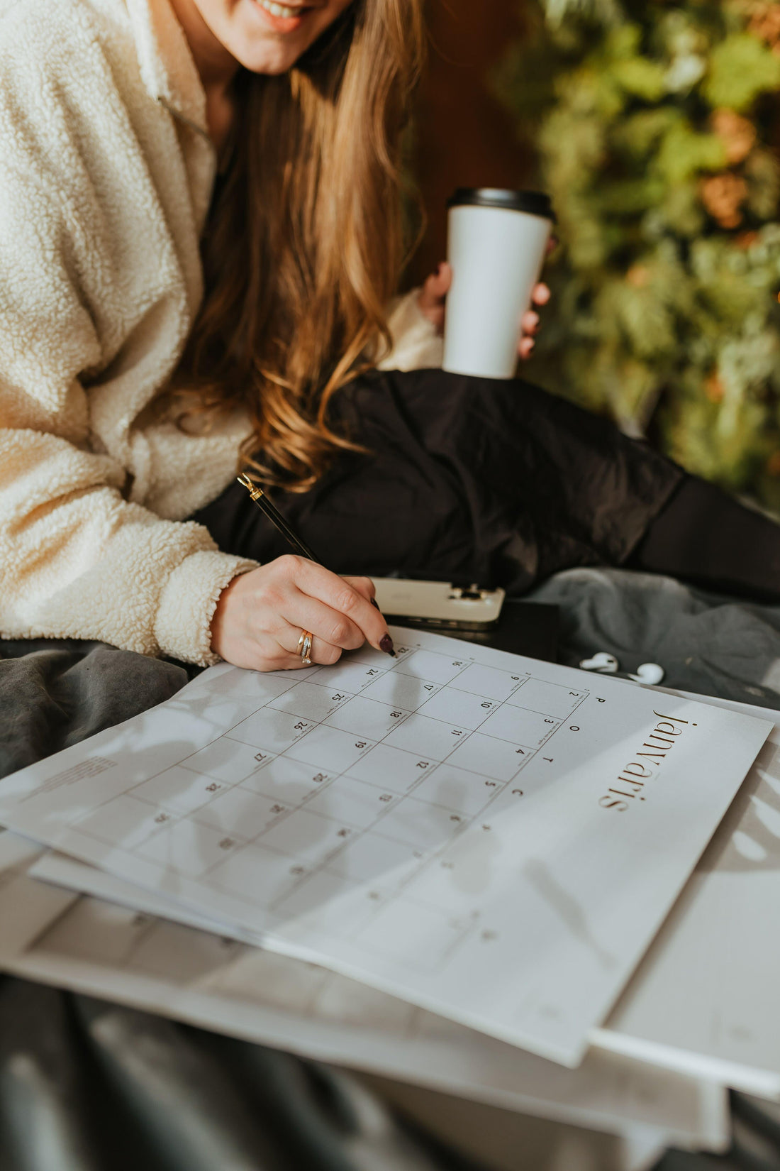Woman in cozy fleece writing on January calendar with coffee cup, relaxing on bed.
