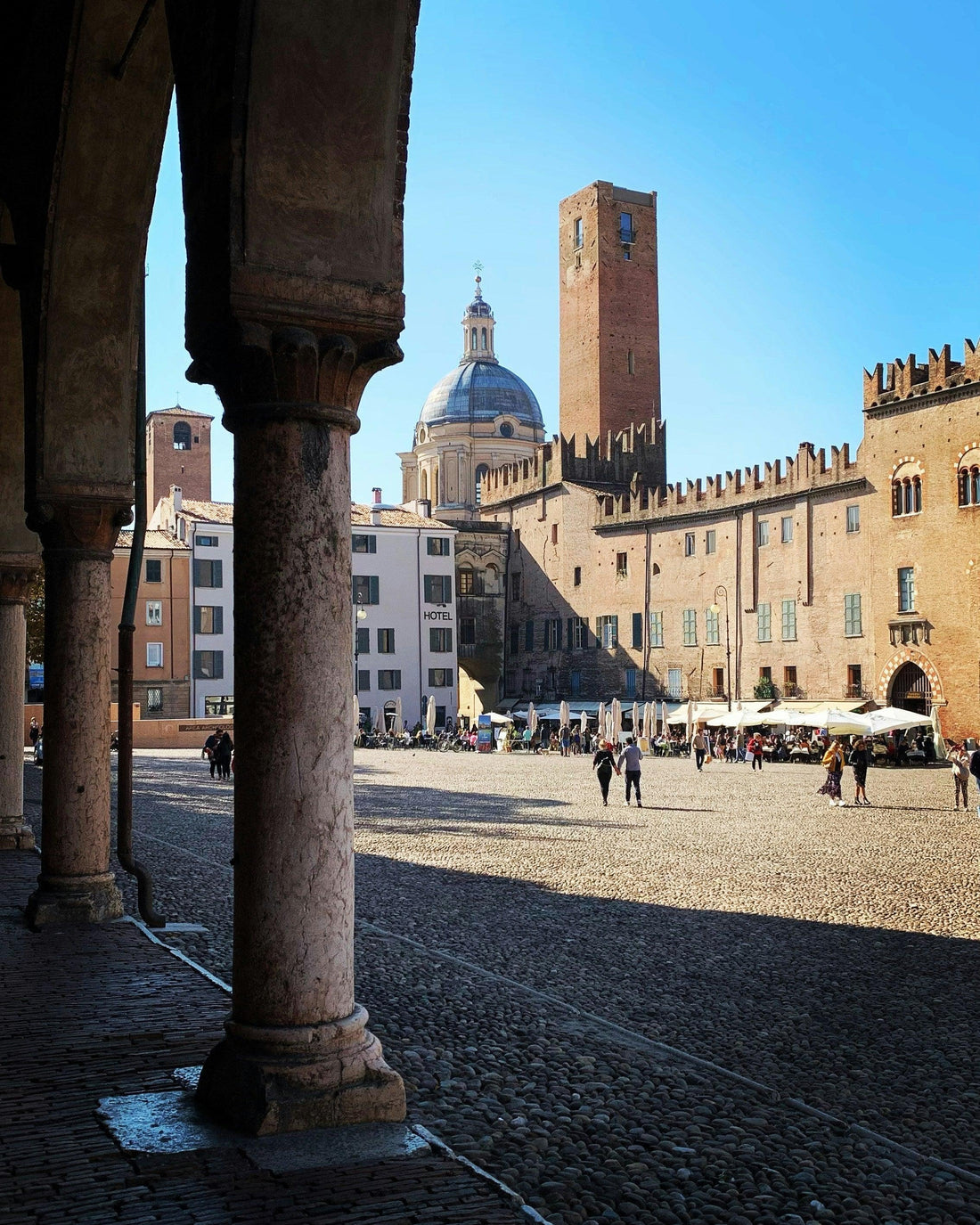 Italian piazza with historic brick buildings, arched columns, and a domed cathedral under blue sky