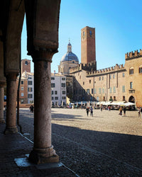 Italian piazza with historic brick buildings, arched columns, and a domed cathedral under blue sky