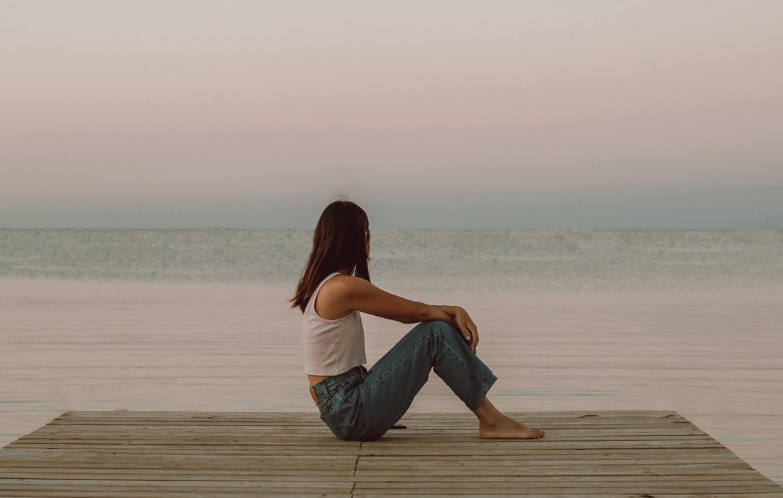 Woman in casual jeans and tank top sitting on wooden pier by calm sea at sunset