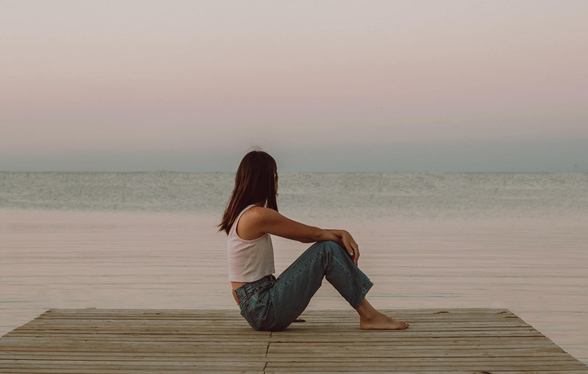 Woman in casual jeans and tank top sitting on wooden pier by calm sea at sunset
