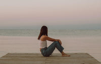 Woman in casual jeans and tank top sitting on wooden pier by calm sea at sunset