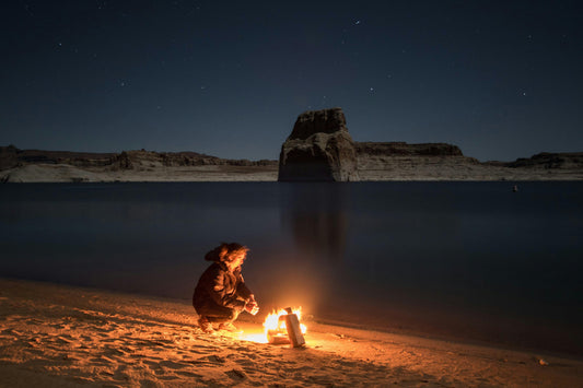 Person sitting by a campfire on a sandy lakeshore at night with rocky cliffs and stars
