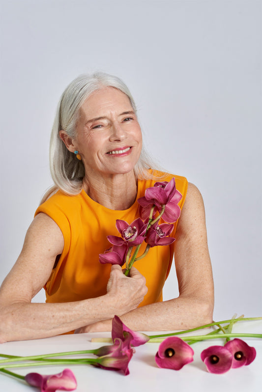 Smiling older woman in orange dress holding purple flowers against a light background.