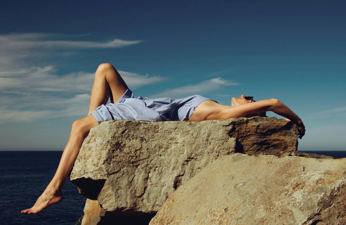 Woman relaxing on sunlit rock by the sea, wearing a light summer dress under blue sky