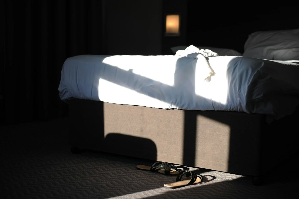 Quiet luxury bedroom scene—white linens in sunlight with sandals under the bed.