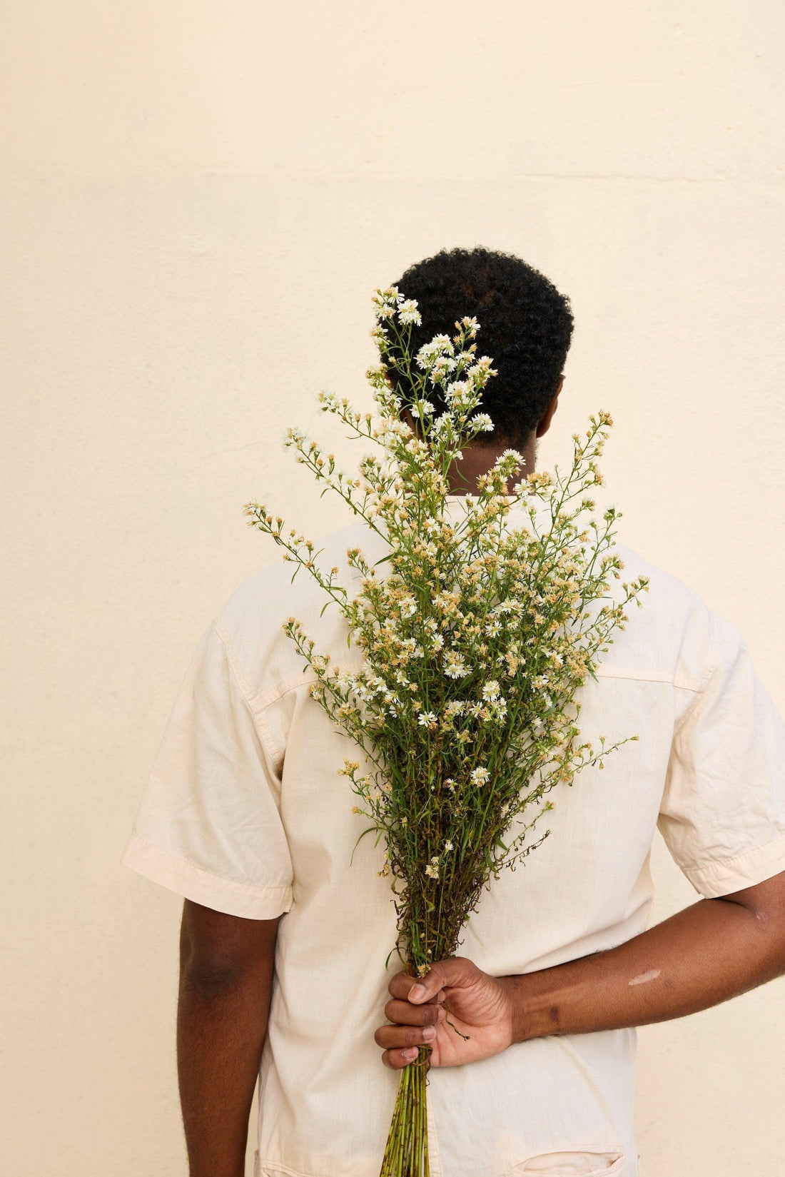 Person in cream shirt holding fresh wildflowers behind back against neutral wall