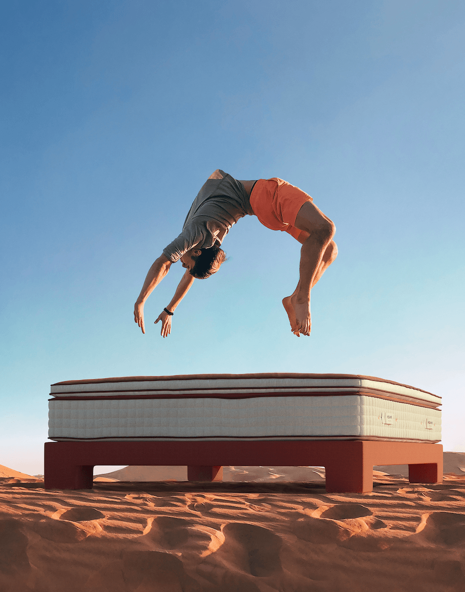 Man doing a backflip above a luxury mattress on a modern bed frame in a desert landscape
