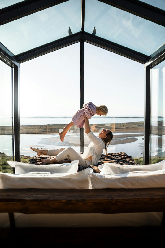 A mother lifts her baby in the air while relaxing on a luxury bed in a glass-walled room.