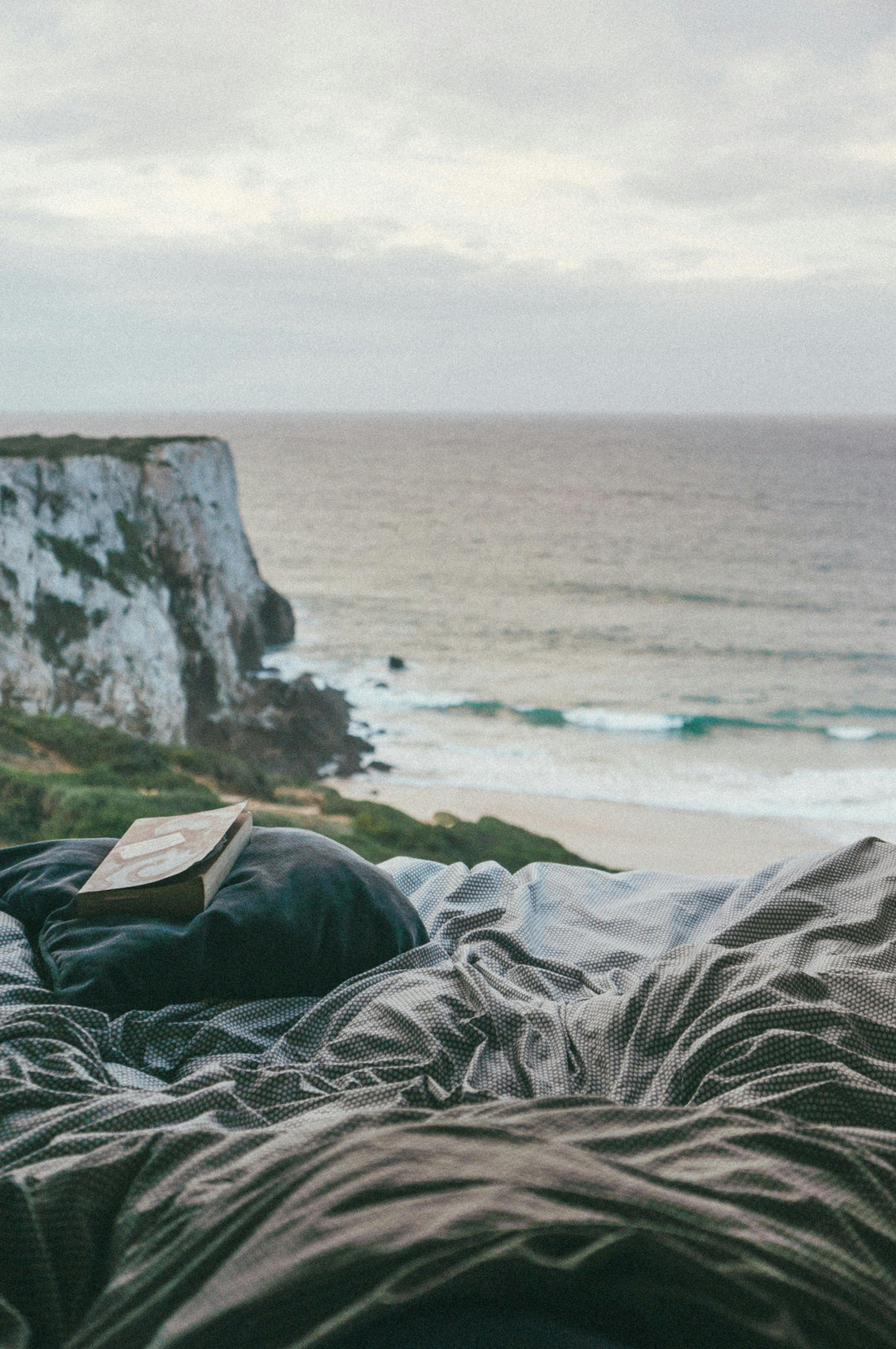 Cozy unmade bed with pillow and book overlooking a seaside cliff and ocean view at sunset